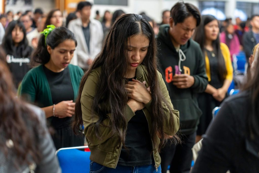 Many individuals of various background are gathered in a large room, engaged in prayer, embodying a spirit of love.